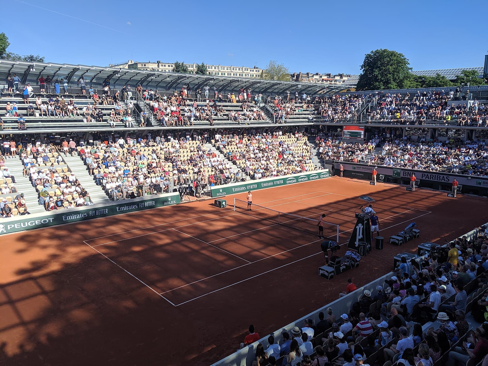 Clay court surrounded by tiered seating and glasshouse architecture at Court Simonne-Mathieu.